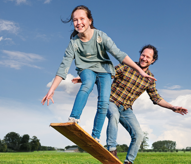 A man and a child jumping on a wooden board in a field.